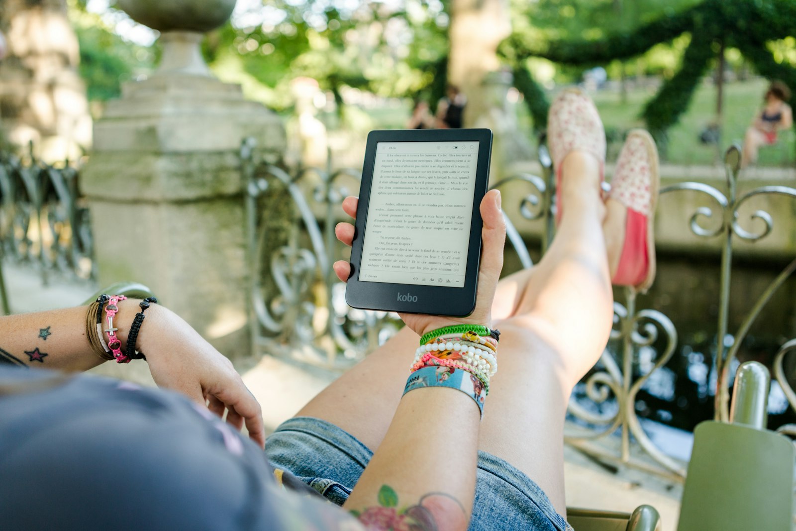 Someone lying back on a park chair in the sun, feet up on the railing, reading on a Kobo
